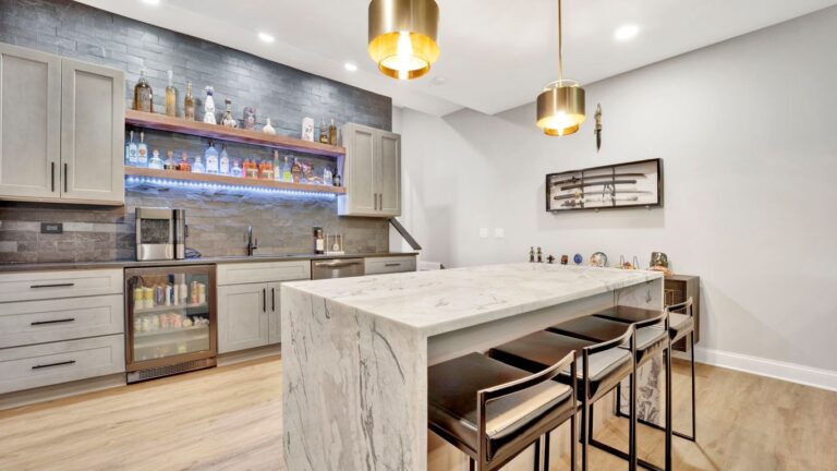 Modern home bar with marble island, bar stools, and liquor display.